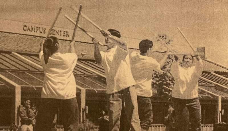 Four people perform a dance with wooden sticks in front of a building labeled Campus Center.