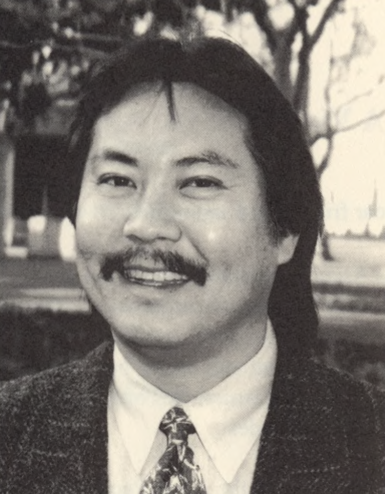A black and white photo of a person with long hair and a mustache, wearing a suit jacket, white shirt, and patterned tie. 