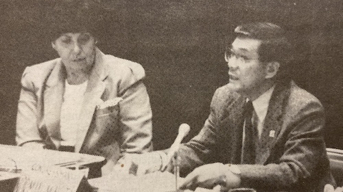 Two people in formal attire are seated at a table with microphones, engaged in discussion. The person on the left wears a suit and the person on the right is speaking while gesturing with one hand. 