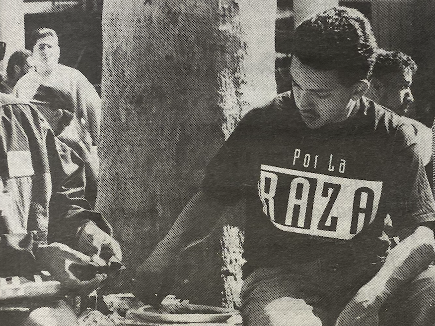 A black and white photo shows a young Latino man sitting near a tree, wearing a T-shirt with Por La Raza printed on it. 