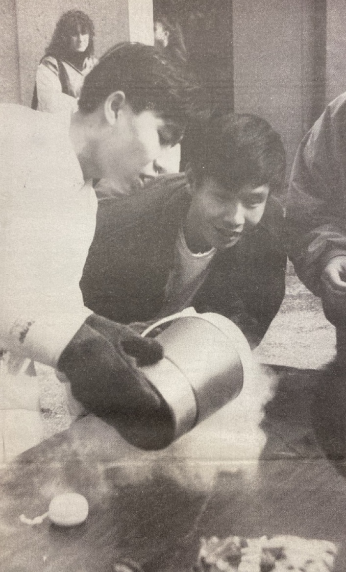 Two Asian students are participating in a science activity. One person is wearing gloves and pouring liquid from a container, creating vapor. The other person watches with interest. A small crowd observes in the background.