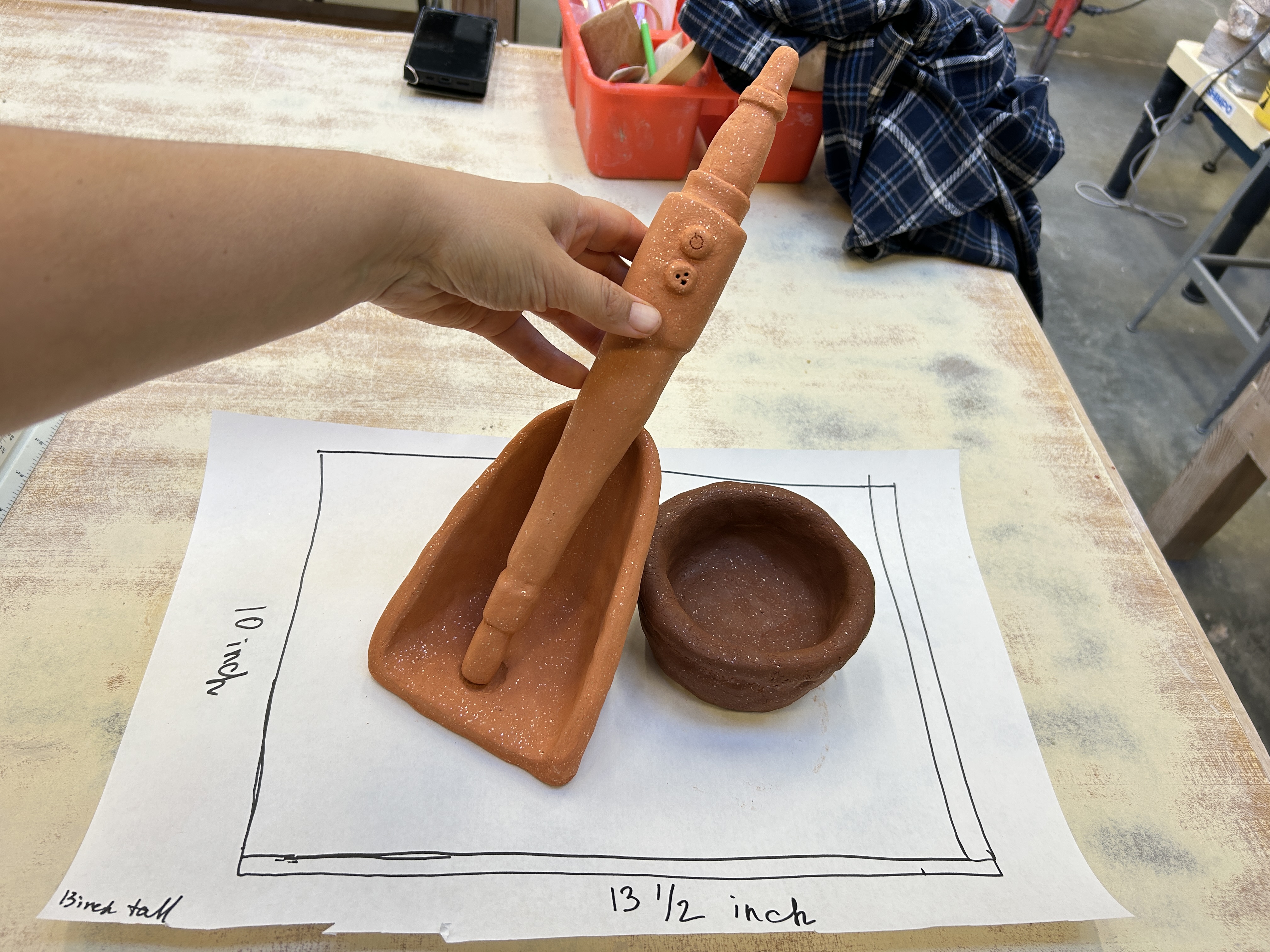 Terracotta clay sculpture of a dustpan with a figural handle, alongside a small round bowl, displayed on a paper template with measurements